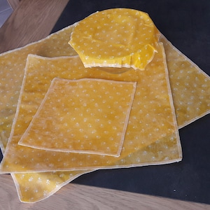 Peut inclure: Quatre emballages alimentaires en cire d'abeille jaune avec un motif floral blanc. Les emballages sont de tailles différentes et sont disposés sur une surface en bois.