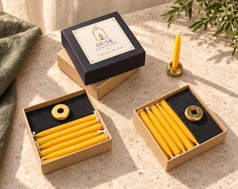 May include: Two open cardboard boxes containing yellow taper candles and a brass candle holder, with a closed black box labelled Dusk Sanctuary A quiet glow after dusk and a single candle in a brass holder nearby.