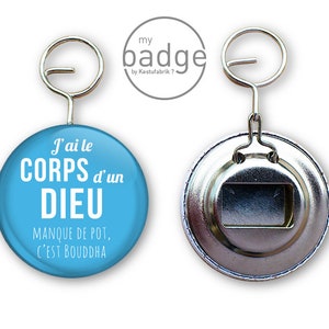 Peut inclure: Un bouton rond bleu et blanc avec le texte "J'ai le CORPS d'un DIEU MANQUE DE POT, C'EST BOUDDHA" dessus. Le bouton est attaché à un ouvre-bouteille argenté.