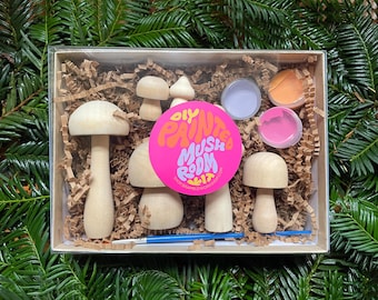 May include: A DIY mushroom painting kit in a clear box. The kit includes several unfinished wooden mushroom figurines, small pots of paint in pink, orange, and purple, a paintbrush, and a sticker that reads "DIY Painted Mushroom Room". The box is filled with brown paper filler.