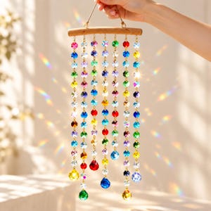 Puede incluir: Un atrapasueños colgante con múltiples hilos verticales de cuentas de vidrio facetadas y multicolores suspendidas de una rama de madera horizontal. Las cuentas presentan varios colores, incluyendo rojo, azul, verde, amarillo y transparente.