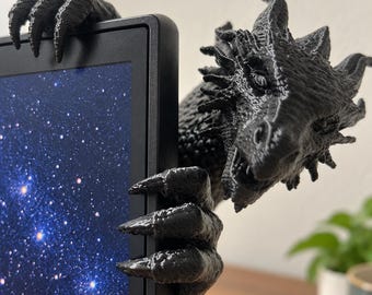 Op de afbeelding: Een zwart 3D-geprint drakenfiguur met getextureerde schubben en scherpe klauwen, ontworpen om aan de rand van een plat beeldscherm te klemmen. De draak heeft een gedetailleerde kop met hoorns en kijkt om de rand van het zwarte frame heen.