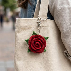 May include: A red crocheted rose keychain with green leaves attached to a silver-toned metal lobster clasp and chain. The floral accessory is secured to the strap of a beige canvas tote bag, showcasing detailed yarn stitching.