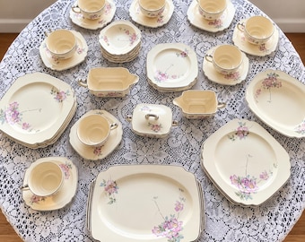 Puede incluir: Un juego de vajilla de porcelana color crema con un delicado patrón floral rosa y morado con tallos verdes. La colección incluye platos cuadrados, platillos, tazas de té, un azucarero con tapa, una salsera y una gran bandeja rectangular.