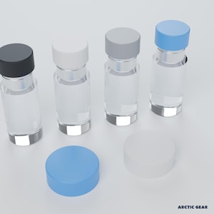 May include: Four clear glass bottles with cylindrical bodies and screw-top lids in black, white, gray, and light blue. Two additional loose lids in light blue and white are placed in the foreground. The text ARCTIC GEAR is visible.
