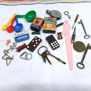 May include: A collection of vintage items including measuring spoons, small boxes, dominoes, keys, a pink plastic knife, and a metal puzzle. The items are arranged on a white surface.