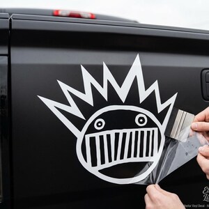 May include: A white vinyl decal featuring a stylized circular face with spiky hair and a wide, toothy grin applied to a matte black truck tailgate. The decal is being smoothed onto the surface using a small, rectangular plastic squeegee tool.