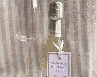 Può includere: Un flûte da champagne in vetro trasparente accanto a una piccola bottiglia di Prosecco. Un nastro bianco lega un'etichetta di carta rettangolare al collo della bottiglia. L'etichetta presenta un bordo marrone smerlato, un'illustrazione di un orso, due calici da champagne e il testo POP & CHEER WHEN BABY IS HERE!