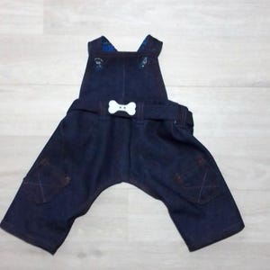 May include: Dark blue denim overalls featuring a white bone-shaped button centered on the waist belt. The garment includes two front pockets with stitched detailing and a blue patterned lining visible at the top of the bib and shoulder straps.
