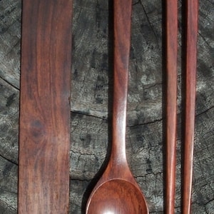 May include: A set of wooden kitchen utensils, including a spoon, a spatula, and a pair of chopsticks. The utensils are made of dark brown wood and have a natural finish.