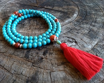 May include: A turquoise blue mala bead necklace with red and silver accents and a red tassel. The necklace is laid out on a wooden surface.