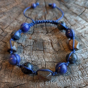 Könnte beinhalten: Ein handgefertigtes Armband mit blauen, schwarzen und braunen Edelsteinperlen. Die Perlen sind auf einer blauen Kordel im Makramee-Stil aufgefädelt. Das Armband ist verstellbar und liegt auf einer Holzoberfläche.