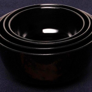 Może przedstawiać: Zestaw czterech czarnych misek do wkładania w siebie. Miski są wykonane z gładkiego, błyszczącego materiału i są ułożone jedna na drugiej.