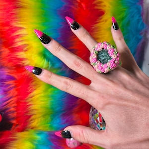 May include: A chunky ring featuring a textured, multi-colored bead-like surface in shades of pink, green, and yellow surrounding a dark, glittery center. The ring is worn on a finger with long, pointed nails painted in a black and pink gradient.