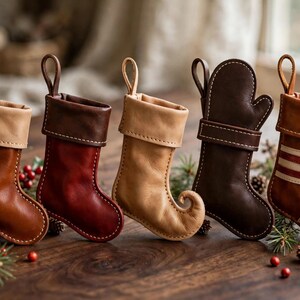 May include: Five small leather ornaments shaped like stockings and a mitten, each featuring a hanging loop. The collection includes various shades of brown, red, and tan, with one stocking displaying horizontal stripes and another with a curled toe.