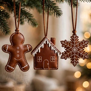 May include: Three brown leather ornaments hanging by thin leather cords. The shapes include a gingerbread person, a house with white stitched roof details, and a snowflake, all featuring white decorative stitching along the edges.