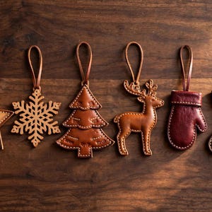 May include: A set of six leather hanging ornaments featuring a star, a snowflake, a Christmas tree, a reindeer, a mitten, and a stocking. Each piece is crafted from brown or dark red leather with visible light-colored stitching and a leather loop.