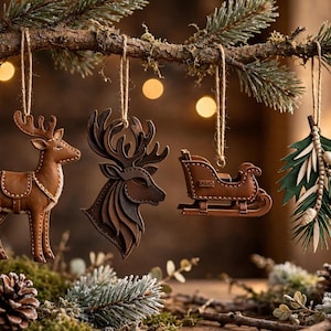May include: Four hanging ornaments suspended from a pine branch: a brown leather reindeer, a layered brown deer head silhouette, a brown leather sleigh, and a green leaf sprig with white berries, all featuring visible stitching details.