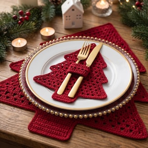May include: A white dinner plate with a gold rim sits atop a red crocheted Christmas tree placemat. A matching red crocheted Christmas tree napkin holder secures a gold fork and knife on the plate. The set features intricate yarn stitching.