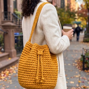 May include: A mustard yellow bucket bag crafted from thick, textured crochet yarn. The bag features a single shoulder strap and a drawstring closure with two dangling knotted ends, providing a secure and structured opening for the main compartment.
