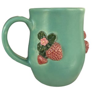 May include: A teal ceramic mug with a curved handle. The mug is decorated with raised strawberry and flower details. The strawberries are red with textured surfaces, and the flowers are pink. The mug has a smooth, glossy finish.