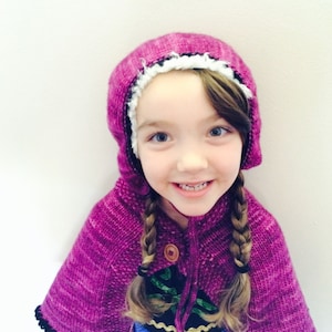 May include: A young girl wearing a pink and white knitted hooded cape with a black trim. She is wearing a blue dress with gold accents and has her hair in two braids.