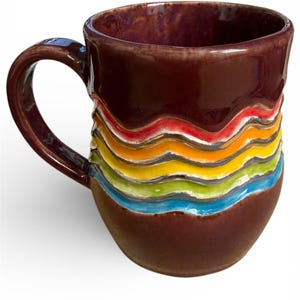 May include: A ceramic coffee mug with a dark brown exterior and a curved handle. The mug features a colorful, wavy design in red, orange, yellow, green, and blue. The interior of the mug is a deep purple color.