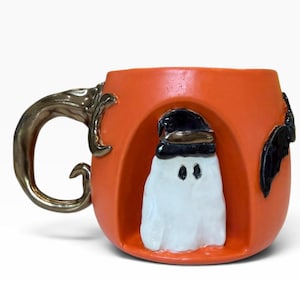 May include: An orange ceramic mug featuring a sculpted white ghost wearing a black hat centered in a recessed arch. The mug has a metallic bronze-colored handle shaped like a curved branch and a black bat figure attached to the side of the body.