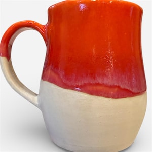 May include: A handmade ceramic mug with a vibrant red glaze on the upper half and a creamy white base. The mug features a curved handle and a smooth, glossy finish, perfect for enjoying hot beverages. The mug's design is simple and elegant.