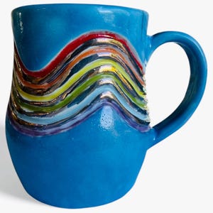 May include: A blue ceramic mug with a colourful, wavy design. The design includes red, orange, yellow, green, and purple, with gold accents. The mug has a curved handle and a smooth, glossy finish. The mug is suitable for hot or cold beverages.
