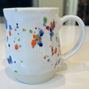 May include: White ceramic mug with a large handle, decorated with colorful paint splatters in blue, orange, green, and red. The mug has a slightly off-white base and a smooth, glossy finish. A handmade, unique coffee mug.