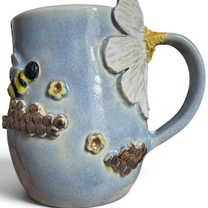 May include: A light blue ceramic mug featuring a three-dimensional sculpted white flower attached to the handle, a small yellow and black bee, and raised metallic bronze honeycomb patterns with tiny yellow flowers scattered across the surface.