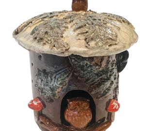 Puede incluir: Una casa de búho de cerámica hecha a mano con un techo en forma de hongo. La casa es marrón con detalles en verde y blanco, con una abertura en forma de búho y detalles de hongos rojos. El techo tiene un diseño de hojas texturizadas.
