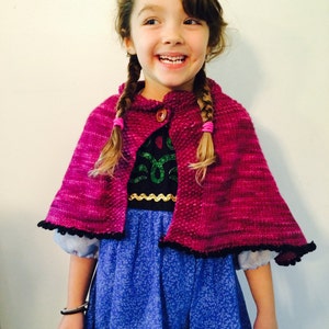 May include: A young girl wearing a pink knitted cape with a black trim and a blue dress with a floral pattern. She is smiling and has her hair in two braids.