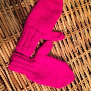 May include: A pair of pink knitted mittens, with one mitten folded over the other, on a woven wicker surface.