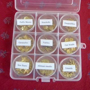 May include: A clear plastic organizer with nine compartments, each holding small, gold-colored charms. Labels identify the charms as "Puffy Moons," "Seashells," "Dragonflies," "Gargoyles," "Fairies," "Cat heads," "Sea Stars," "African masks," and "Lizards."
