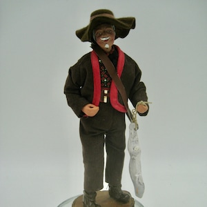 May include: A handcrafted figurine of a man in a brown corduroy suit and hat, holding a white fish. The figure has a painted face and is standing on a wooden base set on a glass circle.