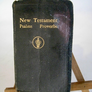 Puede incluir: Un libro negro encuadernado en cuero con letras doradas que dice "Nuevo Testamento Salmos Proverbios". Un emblema dorado de un jarrón está debajo del texto. El libro se exhibe en un pequeño caballete de madera.