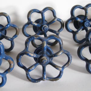 May include: A collection of vintage blue metal valve handles with a distressed finish. The handles have a six-petal flower design and are likely for plumbing or industrial use. The blue paint shows signs of wear, adding to their antique charm.
