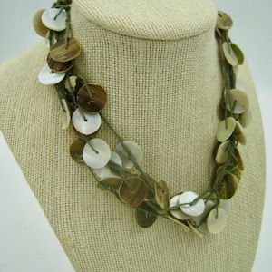 May include: A multi-strand necklace featuring round, flat shell beads in shades of brown, white, and green. The beads are strung on green cord, creating a layered effect. The necklace is displayed on a beige mannequin.