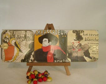 May include: A vintage-style triptych featuring three panels with French advertisements. The central panel showcases a man in a hat and red scarf. The panels are displayed on a small wooden easel, with a cluster of artificial fruit in front.