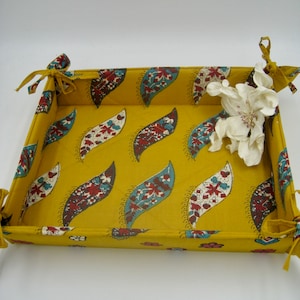 May include: A yellow fabric storage basket with a paisley pattern and white fabric ties. The basket is empty and has a white flower on top.