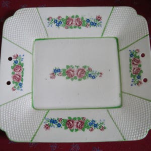 May include: A white, square serving platter with a floral design. The platter features pink roses and blue flowers, with a green border and accents. The edges have a textured pattern and small holes for handles. A vintage piece.