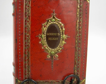 May include: A rectangular red tin box designed to resemble a vintage book, featuring gold-toned metal filigree corners and edges. The center displays an oval plaque with the text "GOURMETS DELIGHT" surrounded by small green and red decorative accents.