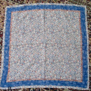 May include: A square pocket square featuring a dense floral pattern in shades of blue, white, and orange. The fabric has a wide, solid blue border with a subtle leaf print, framing the intricate central botanical design on a light background.