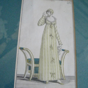 May include: An antique print of a woman in a long, light green and white striped dress with bows, a matching bonnet, and a white collar. She stands near a chair with a teal cushion and white and gold trim. The print has French text.