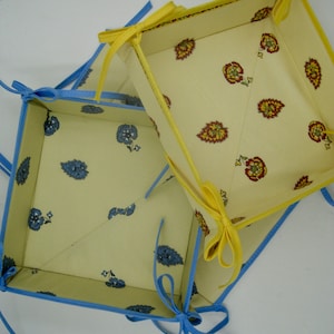 May include: Two fabric storage baskets with blue and yellow trim. The larger basket has a yellow trim and a floral pattern. The smaller basket has a blue trim and a floral pattern.