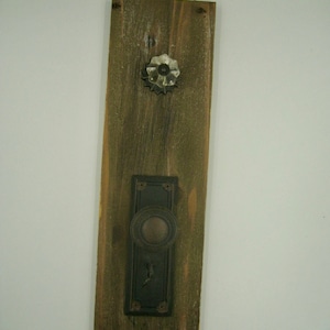 May include: A rustic wooden wall hanging featuring a vintage door knob and backplate. The wooden plank is stained with a weathered finish. A clear glass knob and a metal door handle are attached to the wood.