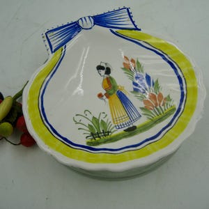May include: A white, scallop-edged ceramic dish with a painted design. The design features a woman in traditional clothing, flowers, and a yellow and blue border. A blue bow detail is at the top. Decorative berries are to the left.