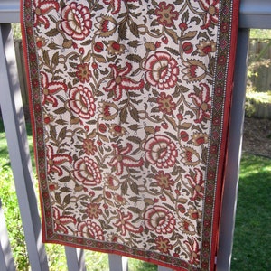 May include: A white table runner with a floral pattern in shades of brown and red. The runner has a red border with a geometric pattern.